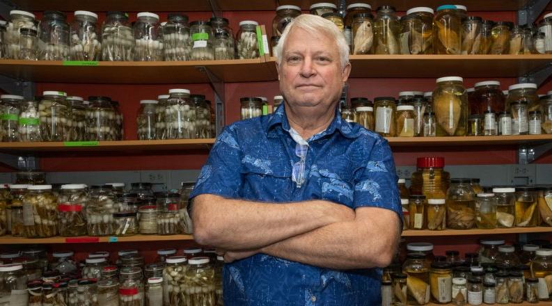 A man in a blue shirt poses in front of jars of biological samples.